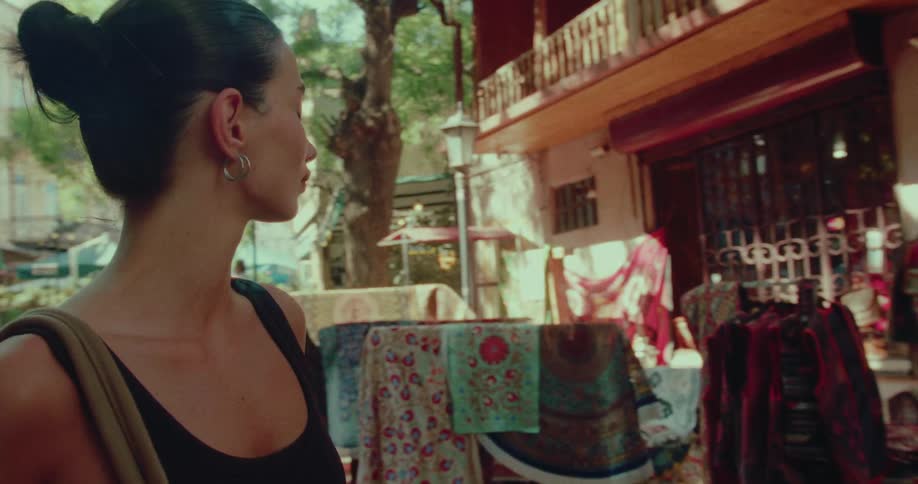 A curious woman wanders through a sunlit street market, surrounded by a vibrant display of traditional textiles and carpets