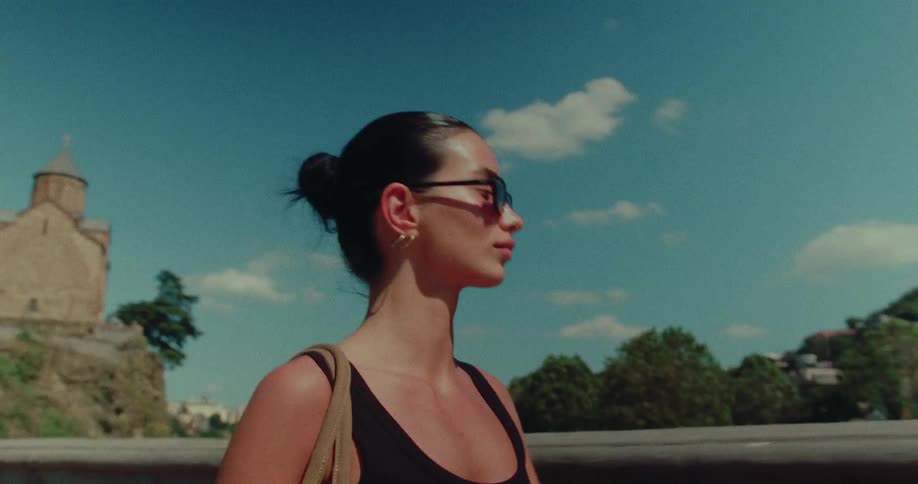 A stylish woman in sunglasses walks across a city bridge on a sunny day, exploring the historic architecture of a European city