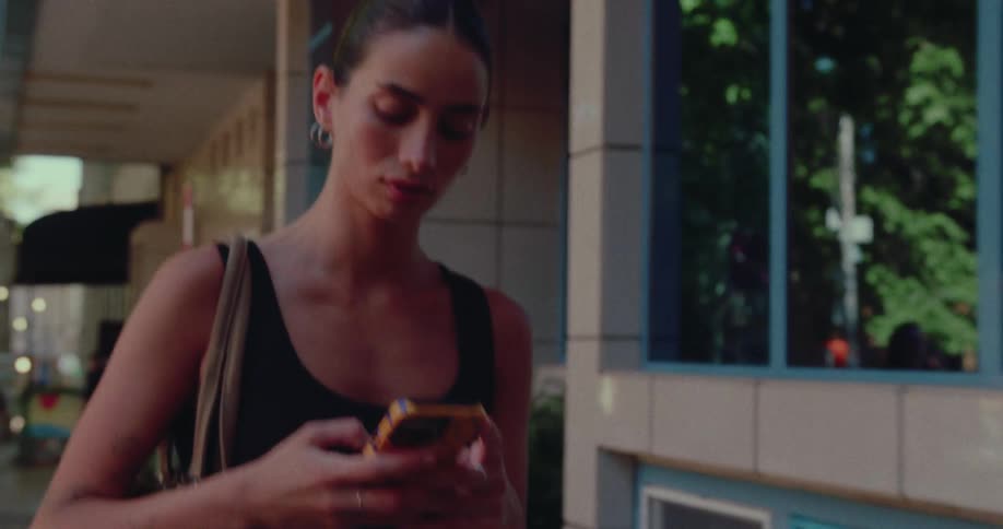 A young woman in a black tank top walks down a city street while intently looking at her smartphone