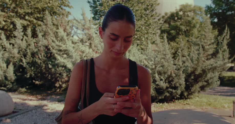A young woman sits on a stone bench in a sunny park, intently scrolling on her smartphone as the camera slowly zooms in