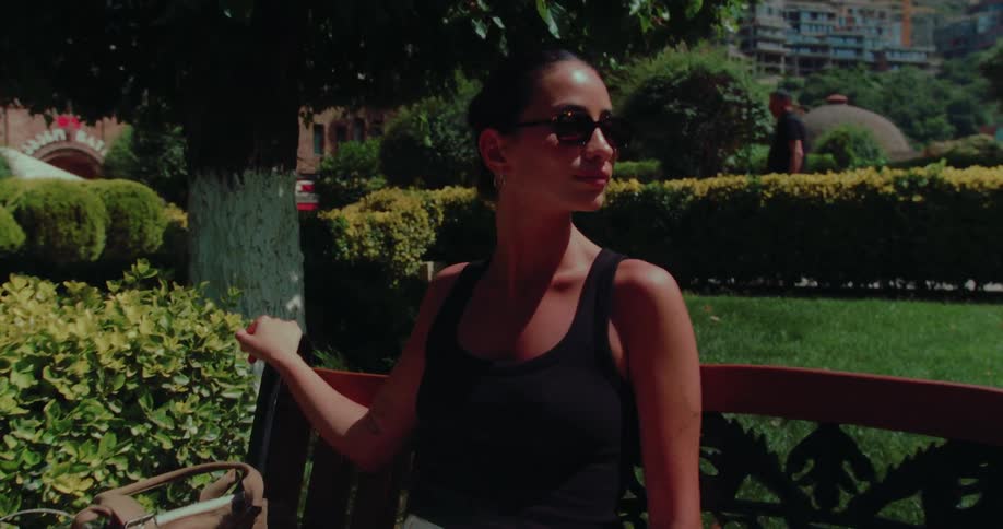 A stylish woman in sunglasses relaxes on a park bench under the shade of a tree on a sunny day