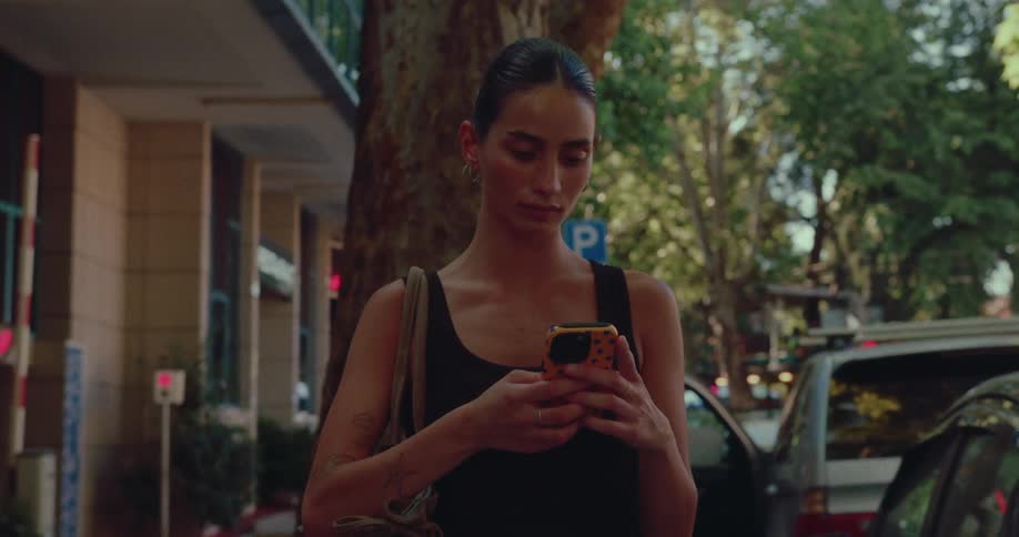 A stylish young woman with slicked-back hair and tattoos stands on a city street, focused on her smartphone