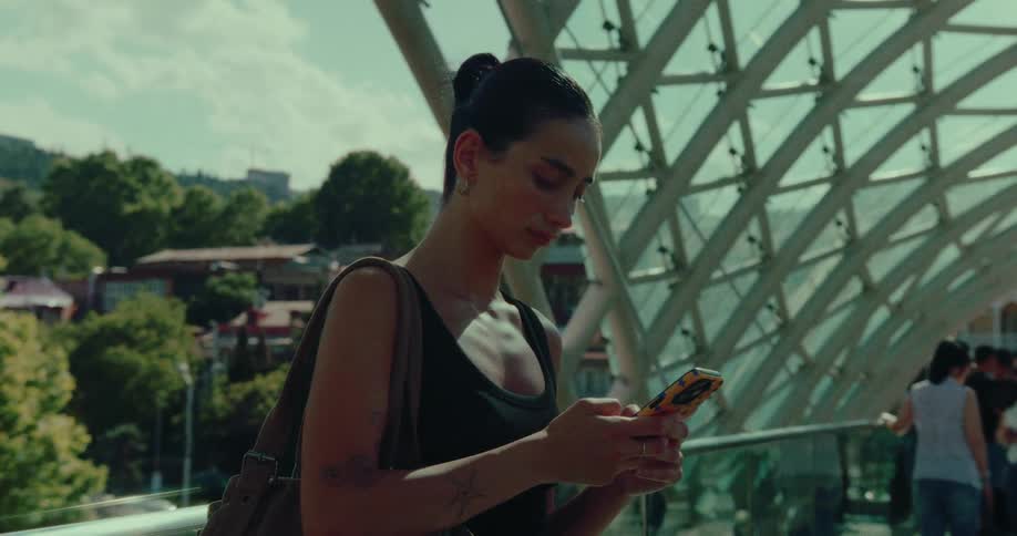 A young woman in a black tank top stands on a modern glass bridge, engrossed in her smartphone on a sunny day