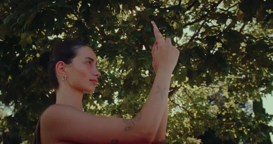 A stylish young woman with her phone raised photographs the sun filtering through the green leaves of a tree in a summer park
