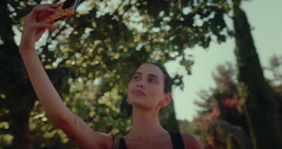A beautiful young woman with her hair pulled back takes a selfie video with her smartphone while enjoying a sunny day in the park