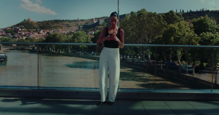 A woman stands on a modern city bridge overlooking a river, absorbed in her smartphone as pedestrians walk by