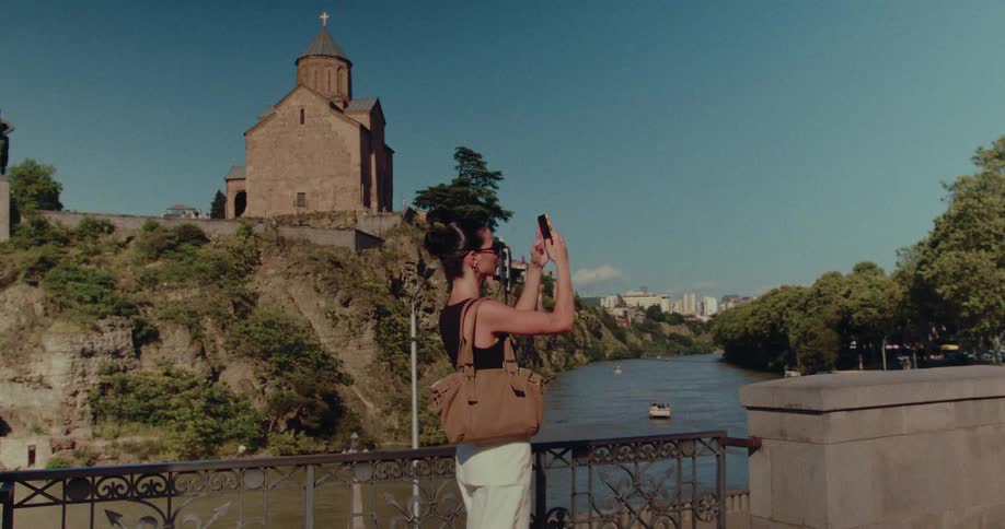 A young woman stands on a bridge and takes a photo of an ancient church overlooking the river