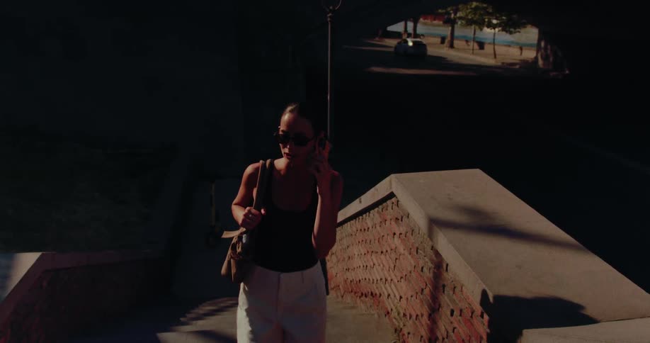A stylish woman in sunglasses walks up urban stairs out of the shadows while talking on her phone