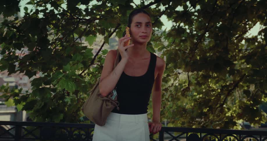A young woman stands on a scenic balcony, answers a call on her smartphone, and engages in a relaxed conversation amidst green foliage