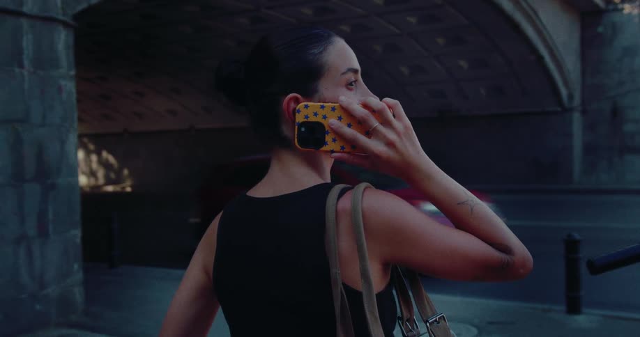 A woman with her hair in a bun talks on her phone while standing on a sidewalk next to a city underpass as traffic passes by