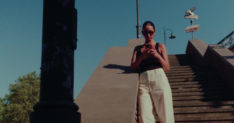 A stylish woman in sunglasses scrolls on her phone while leaning on a concrete barrier at the top of a city staircase on a sunny day