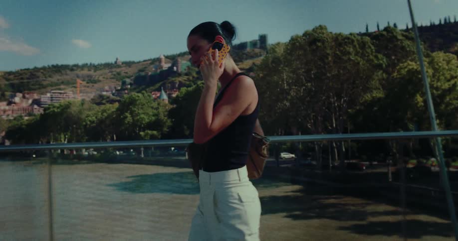 A woman walks along a glass-railed bridge over a river while talking on her phone, with a scenic city on a hill in the background
