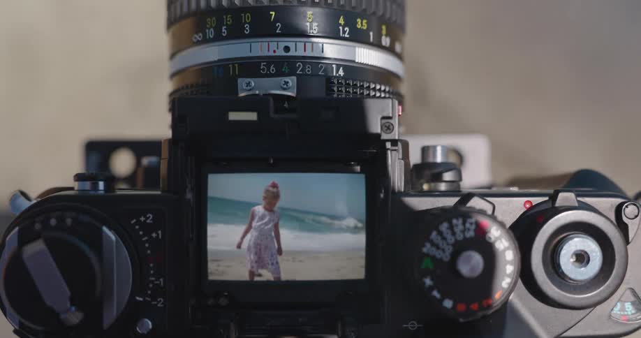 A little girl in a floral dress poses on a sunny beach, seen through the viewfinder of a vintage film camera