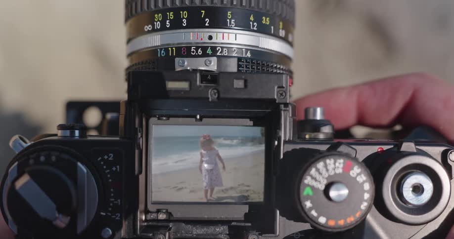 A photographer looks through the viewfinder of a vintage film camera to capture a portrait of a little girl on the beach