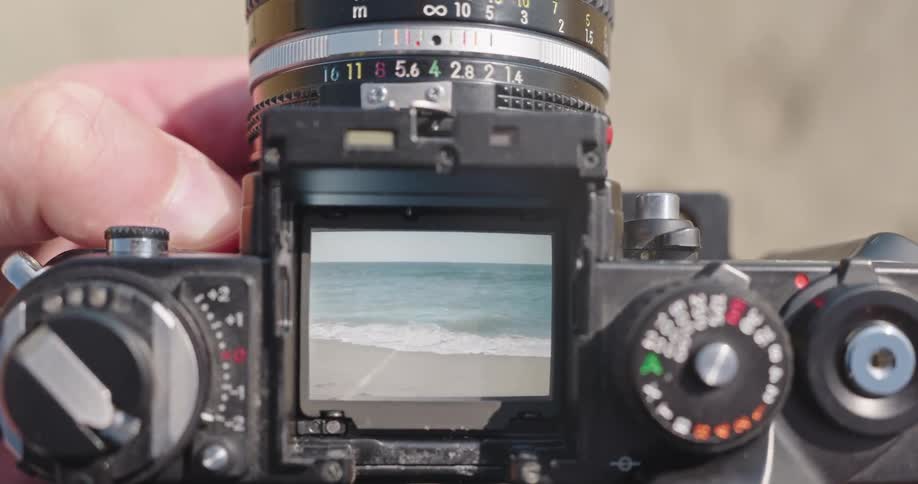 A top-down view looking through the waist-level viewfinder of a vintage SLR camera, focusing on the ocean waves rolling onto the beach