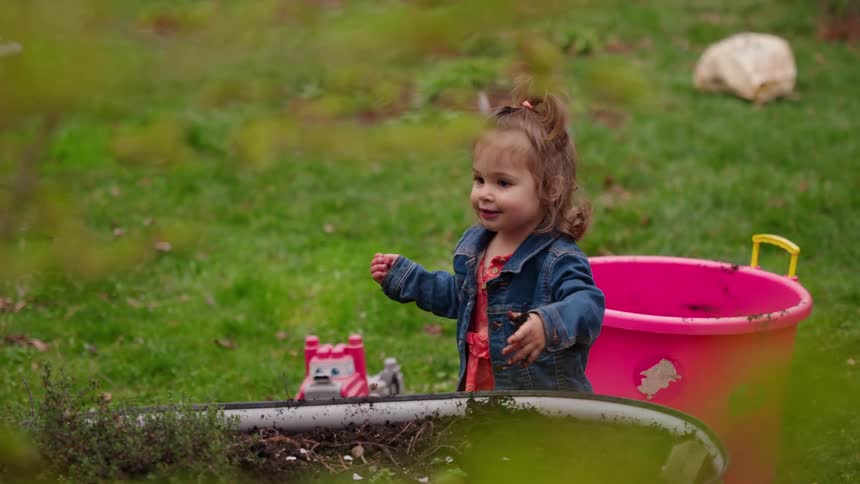 A smiling toddler girl happily plays with the soil in a backyard garden bed in slow motion