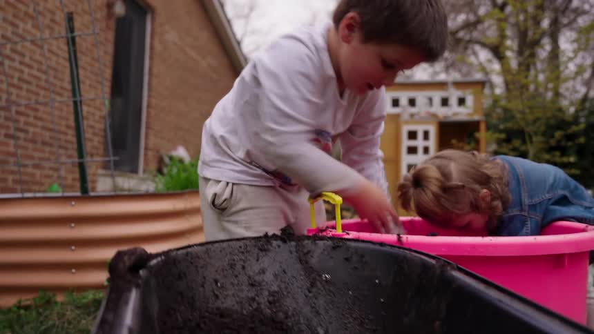 Two adorable siblings get messy while joyfully playing together in the garden, scooping handfuls of dirt into a wheelbarrow