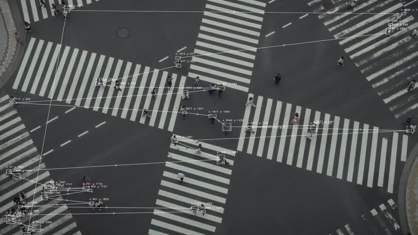 An aerial view shows AI-powered surveillance tracking individuals and their movements across a busy urban crosswalk with data overlays