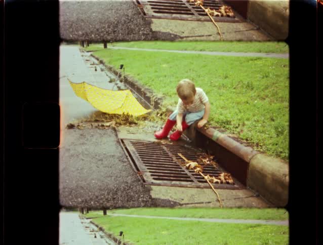 A curious toddler in bright red rain boots sits on the curb and plays by a storm drain after a rain shower