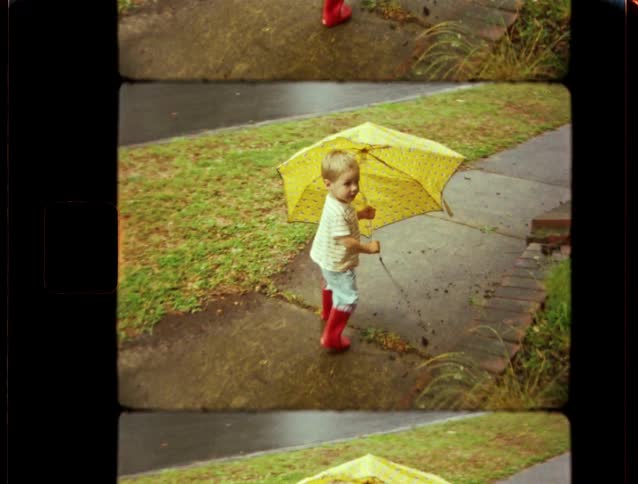 A young boy in red rain boots walks down a wet sidewalk holding a bright yellow umbrella in this vintage-style footage