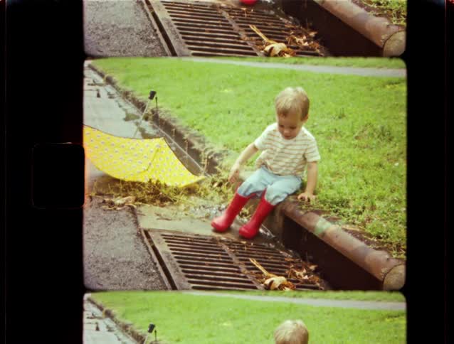 A toddler in bright red rain boots sits on a curb, fascinated by the rainwater flowing into a storm drain with his yellow umbrella nearby