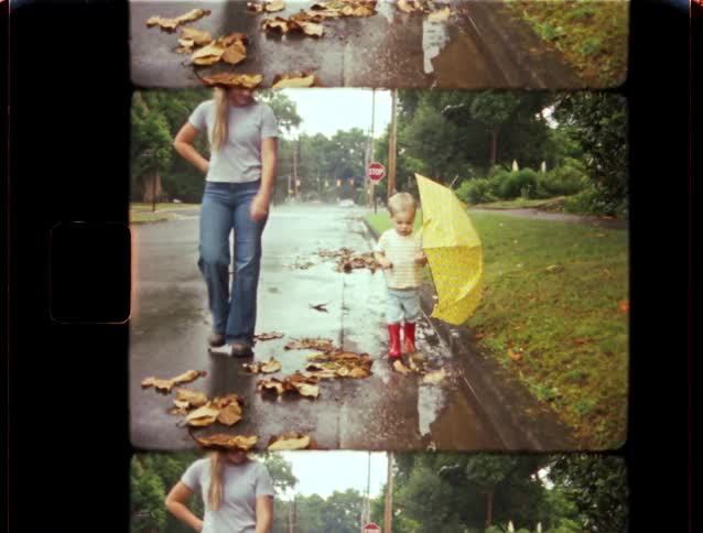 A mother watches as her young son in bright red rain boots splashes in puddles along a suburban street with a yellow umbrella
