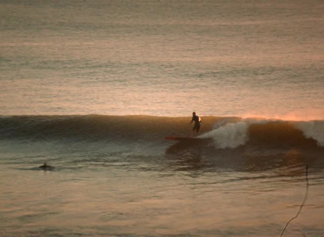 A silhouetted surfer rides a long, gentle wave across the ocean during a golden sunset