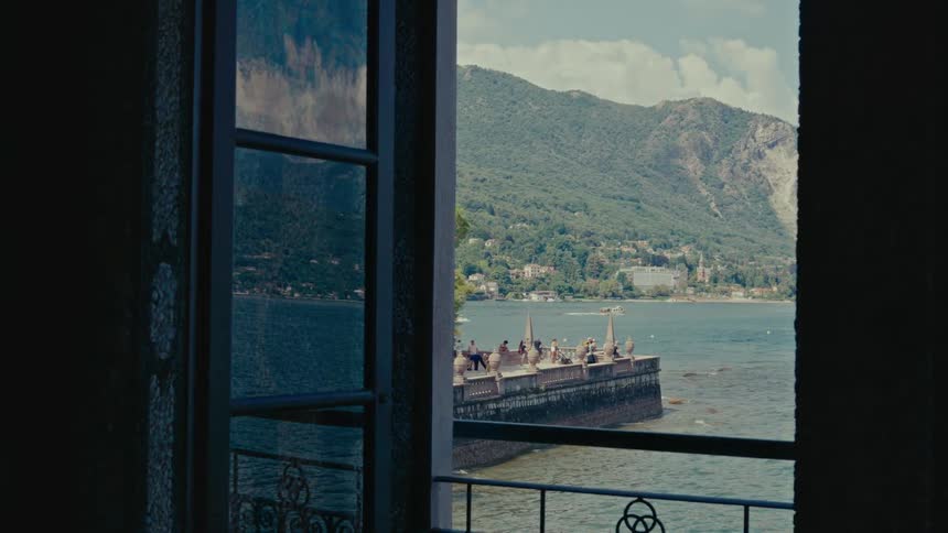A picturesque view from an open window reveals a serene lake, majestic green mountains, and people enjoying a sunny day on a waterside promenade