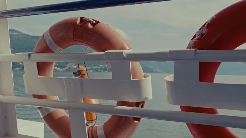 A close-up of an orange lifebuoy on the railing of a boat sailing on a calm lake with a distant shoreline visible