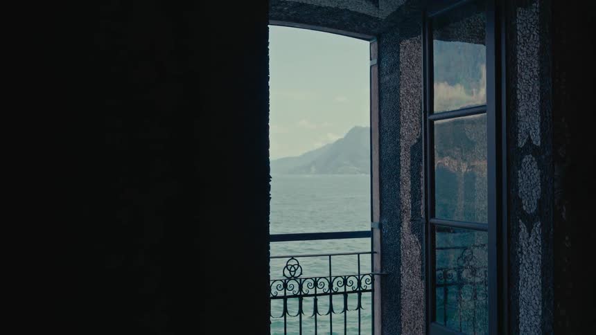 From the shadows of a room, an open window frames a tranquil view of a majestic lake and distant mountains