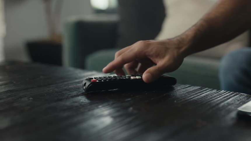 A man's hand picks up a TV remote control from a wooden coffee table in slow motion