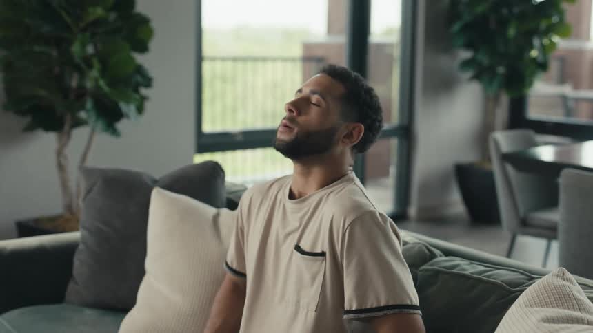 A young man walks into his modern apartment, sits on the couch with a happy sigh of relief, and starts using his smartphone