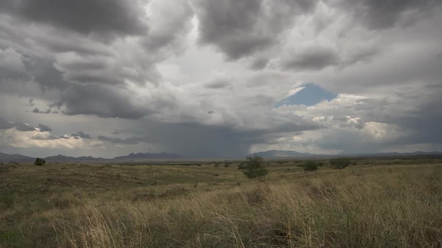 Wind sweeps through the tall grass of a vast prairie as a dramatic storm cell unleashes rain on the distant mountains