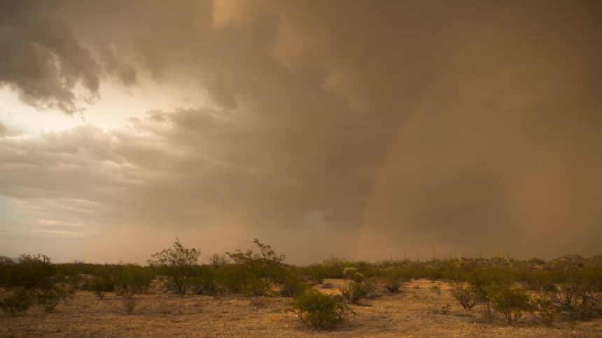 A dramatic time-lapse shows a powerful rainstorm and a vibrant rainbow appearing under dark storm clouds in the desert