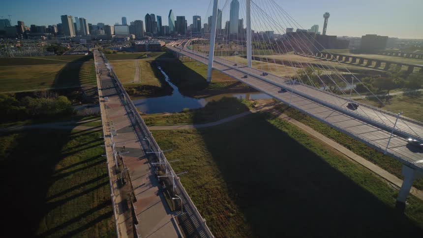 Aerial flight over the Ronald Kirk pedestrian bridge towards the Margaret Hunt Hill Bridge, revealing the Dallas city skyline on a bright, sunny morning