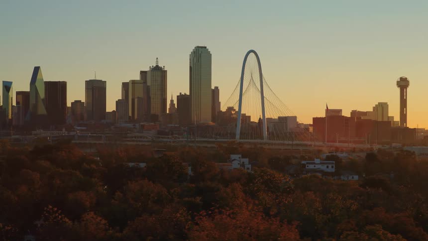 A slow aerial push reveals the iconic Dallas, Texas skyline, including the Margaret Hunt Hill Bridge and Reunion Tower, glowing warmly at sunrise