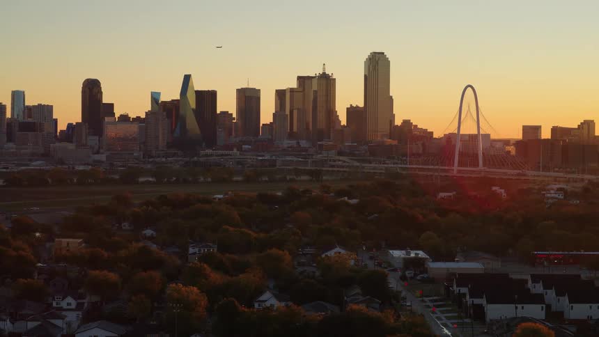 A slow zoom aerial view of the Dallas, Texas skyline with the Margaret Hunt Hill Bridge glowing at sunset as a plane flies by