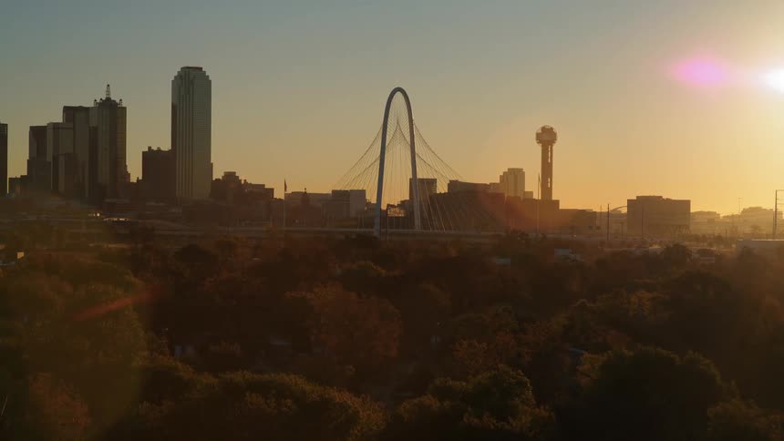 Aerial drone footage pushes over trees to reveal the Dallas, Texas skyline with the Margaret Hunt Hill Bridge at sunrise