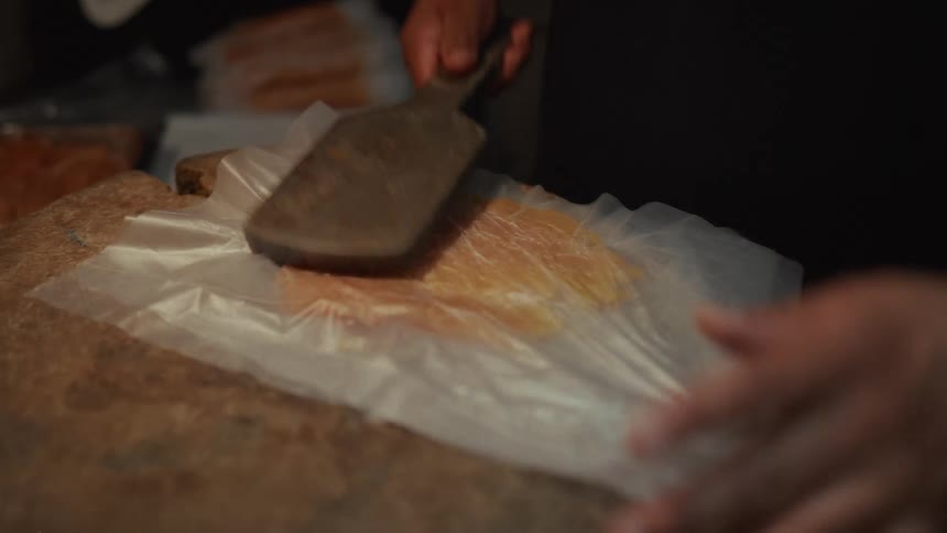 A butcher expertly flattens a thin slice of chicken with a cleaver on a rustic wooden block in slow motion