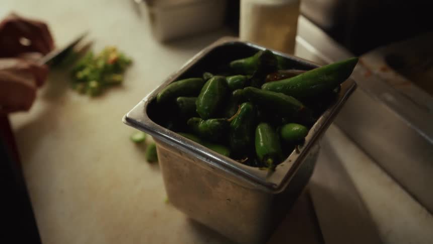 A chef chops fresh jalapeños on a cutting board next to a container full of whole peppers in a professional kitchen