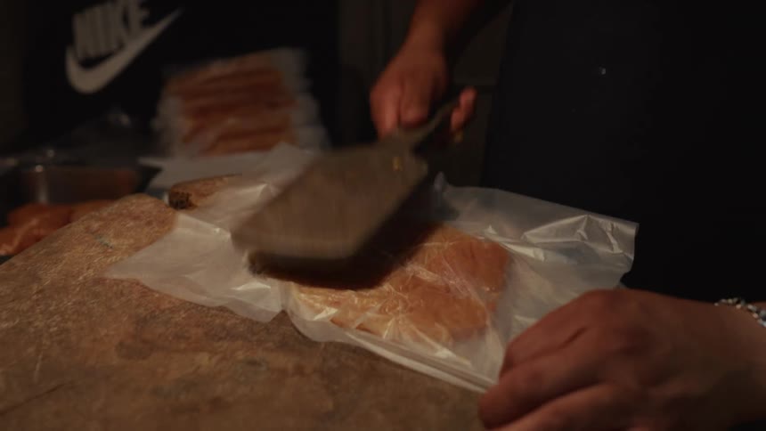 A chef's hands use a meat tenderizer to flatten a chicken breast on a wooden butcher block