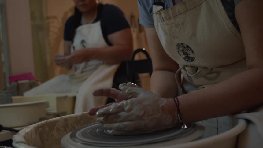 A woman wearing an apron concentrates as she molds wet clay on a spinning pottery wheel in a softly lit studio