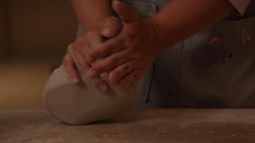 An artisan potter's hands expertly knead and wedge a piece of clay on a studio workbench before sculpting