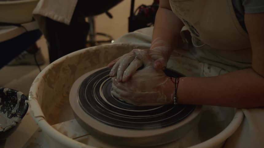 A close-up of a potter's hands covered in wet clay, skillfully centering a lump on a spinning wheel in a workshop