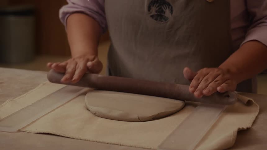 Slow motion close up of a potter's hands using a wooden rolling pin to flatten a piece of clay on a canvas surface