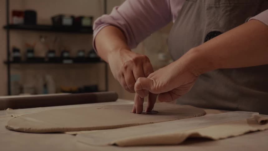 A potter's hands carefully use tools to cut a precise shape from a flattened slab of clay in a studio