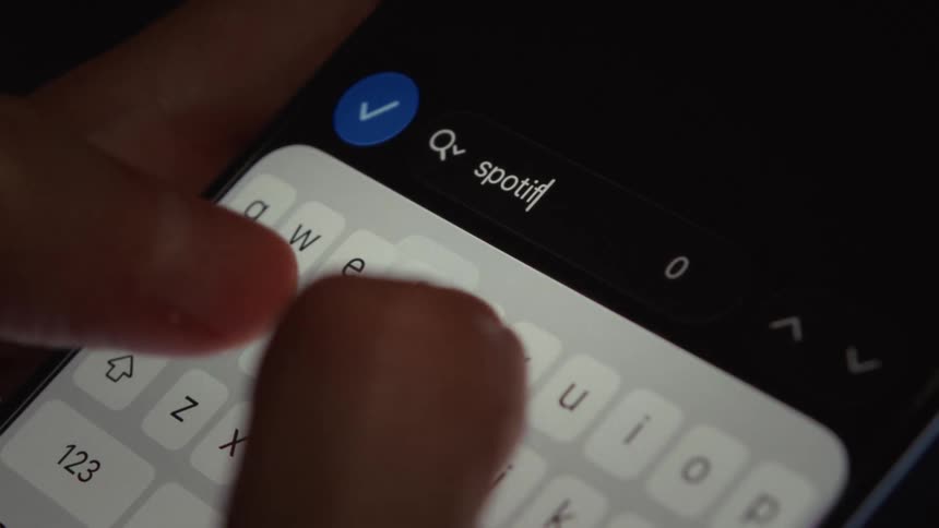 A close-up of fingers typing 'Spotify' into a search bar on a smartphone with a dark mode interface
