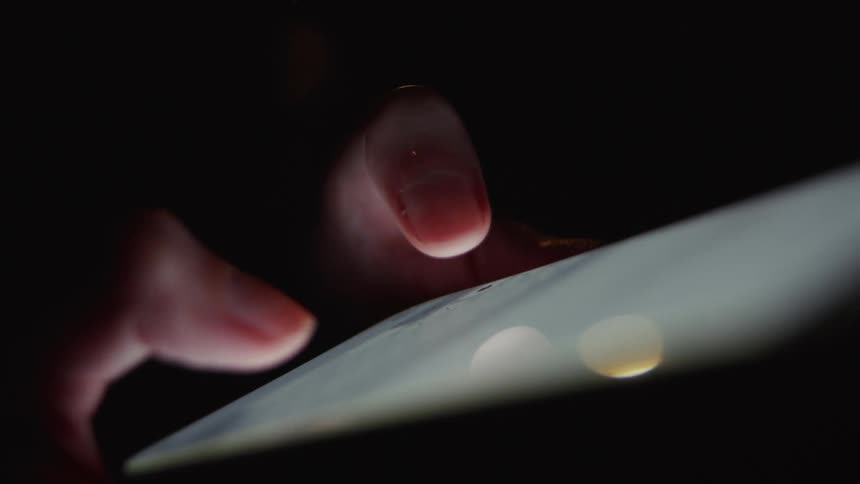 A dramatic close-up of fingers scrolling and tapping on a brightly lit touchscreen in the dark
