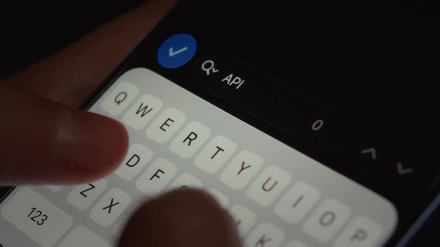 A close-up shot of a finger typing 'API' into a smartphone's search bar in a dimly lit environment