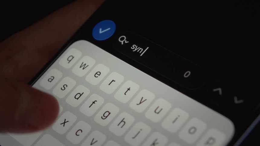 A person's fingers type the word 'sync' into the search bar of a smartphone in dark mode